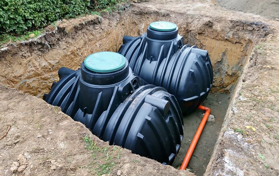 Image shows two black plastic septic tanks installed in a dug-out pit, part of a cesspool services Suffolk County setup. Each tank has a green cover on top. An orange pipe at the bottom connects the tanks. The surrounding soil is freshly excavated, and green foliage is visible in the background.