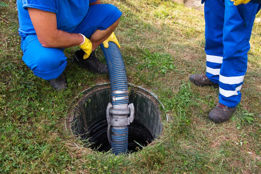 Two workers dressed in blue uniforms and yellow gloves are seen managing a large hose inserted into a sewer manhole on a grassy area. One worker is crouching while the other stands near the manhole, illustrating efficient septic tank cleaning services in Suffolk County.