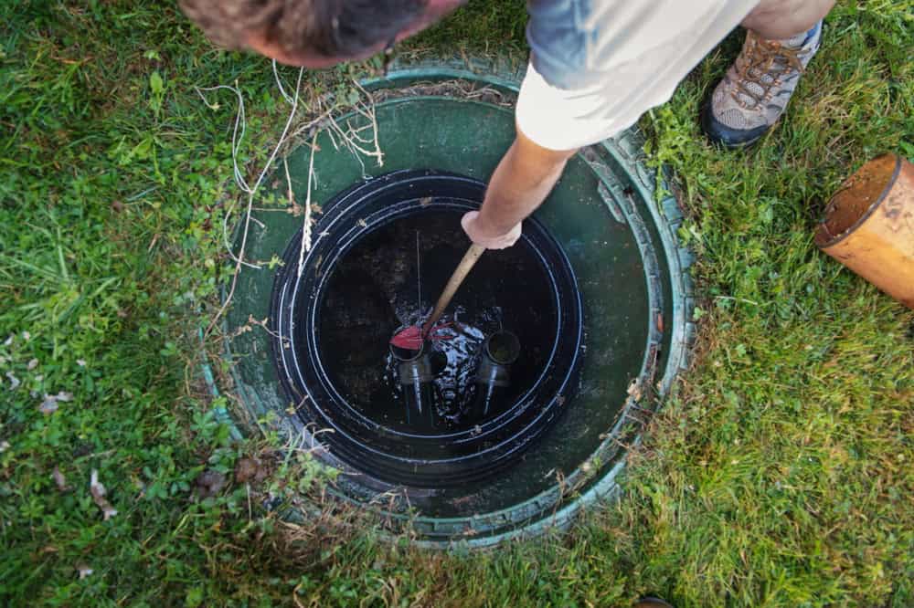 A person is shown from above inspecting or cleaning a septic tank in a grassy area in Suffolk County. They are using a tool to reach inside the open tank, which has a circular cover. A section of the person's head, arm, and boot is visible in the image.