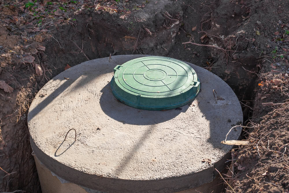 A large, partially buried concrete septic tank with a green circular lid is positioned in an excavated dirt area.