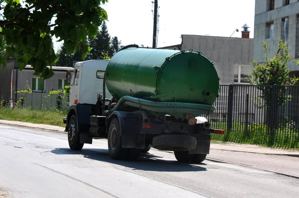 A large green sewage truck drives down a residential street on a sunny day, offering essential cesspool services in Suffolk County. The truck, equipped with a cylindrical tank and visible hoses at the back, moves past trees, buildings, and a fenced property in the background.