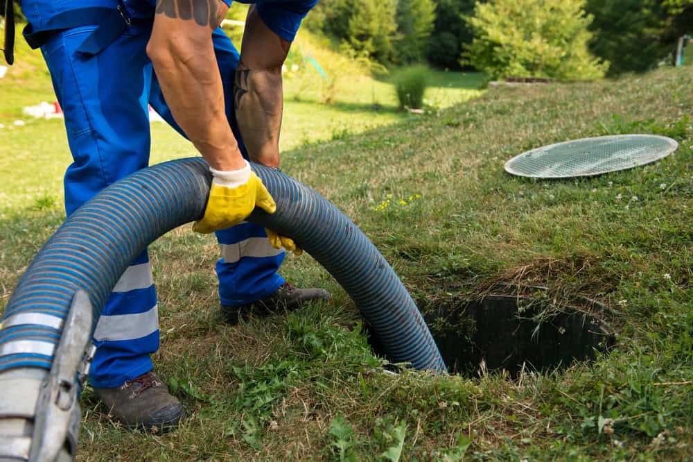 A worker in blue overalls and yellow gloves is holding a large flexible hose, inserting it into an underground septic tank opening on a grassy lawn.