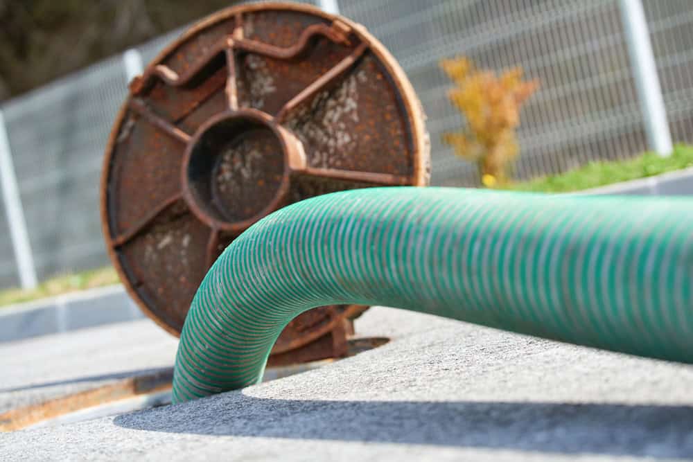 Close-up view of a coiled green hose attached to a rusty metal reel on a paved surface, with a blurred fence and vegetation in the background. The hose bends and inserts into an opening in the ground, suggesting septic tank pumping is underway.