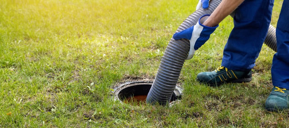 A person in blue work gear and gloves holds a large hose inserted into an open septic tank in a grassy area, clearly indicating septic tank pumping or cleaning.