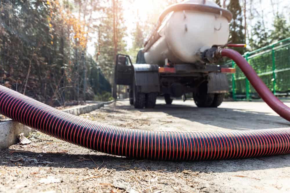 A large tanker truck, used for septic tank pumping, is parked on a paved surface surrounded by trees. A red and black hose extends from the truck, resting on the ground. The sun shines brightly through the trees, casting light and shadows on the scene.