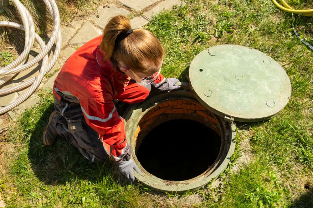 A person in protective clothing and gloves opens a round green manhole cover outdoors, revealing a dark underground space—illustrating the importance of cesspool services Suffolk County, NY. Grass, a stone path, and coiled hoses are visible nearby.