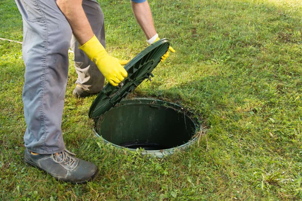 A person wearing yellow gloves and gray pants is lifting the lid off a septic tank access point on a grassy area.