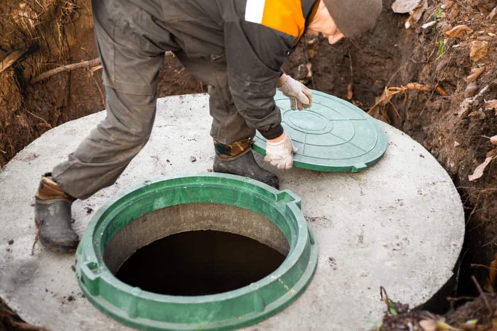 A worker in gloves and work clothes is lifting a green lid off a round concrete septic tank opening outdoors, surrounded by soil and leaves—typical of septic tank services Suffolk County, NY.