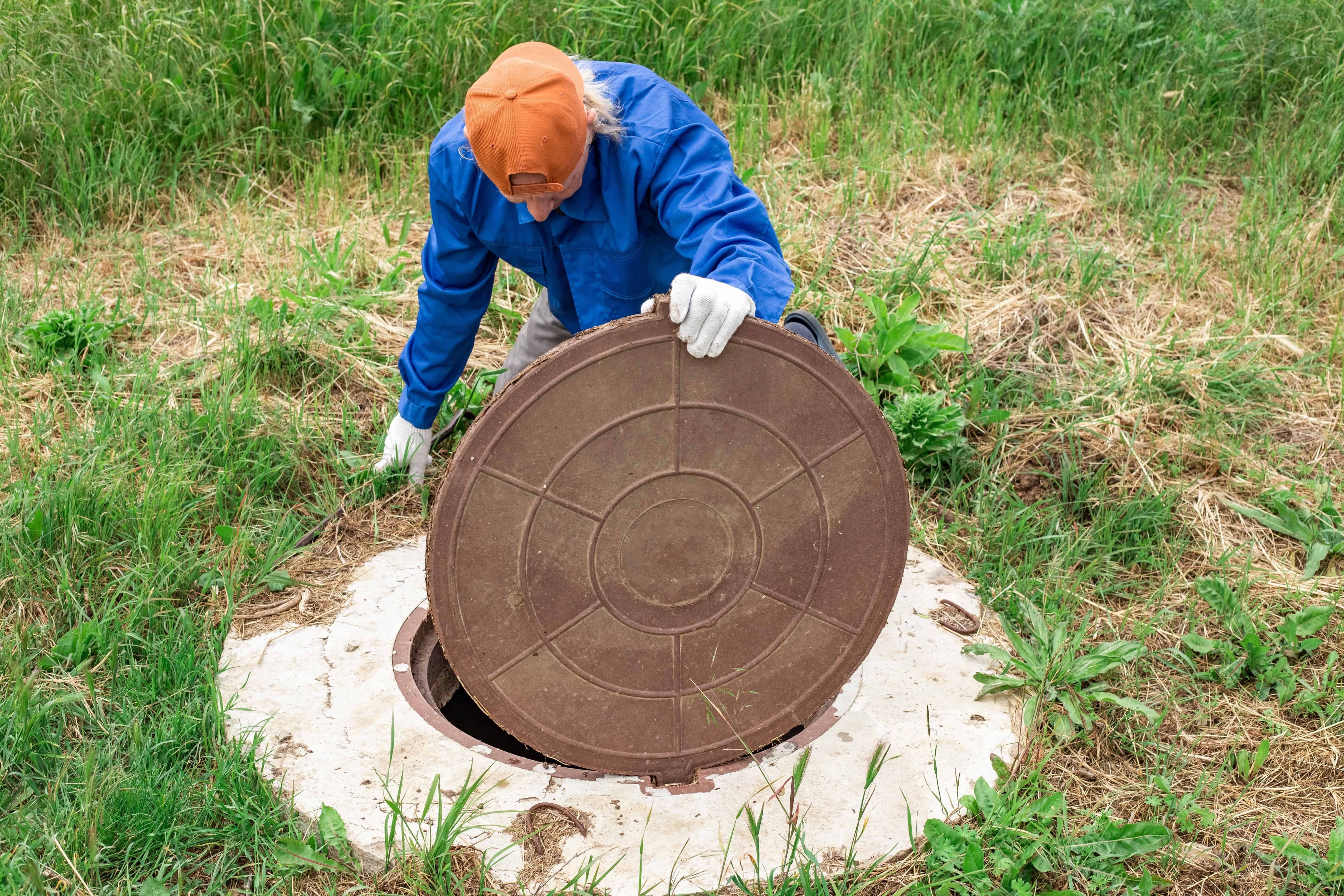 A person wearing an orange cap, blue shirt, and white gloves lifts a large, round manhole cover in a grassy, overgrown area. The individual is bending over while performing the septic tank cleaning task.
