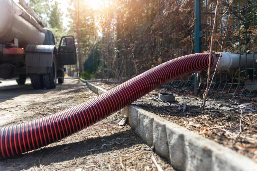 A large red and black hose connected to a NY cesspool services Suffolk County, NY truck is laid on the ground near a curb, with sunlight filtering through trees and a metal fence in the background.
