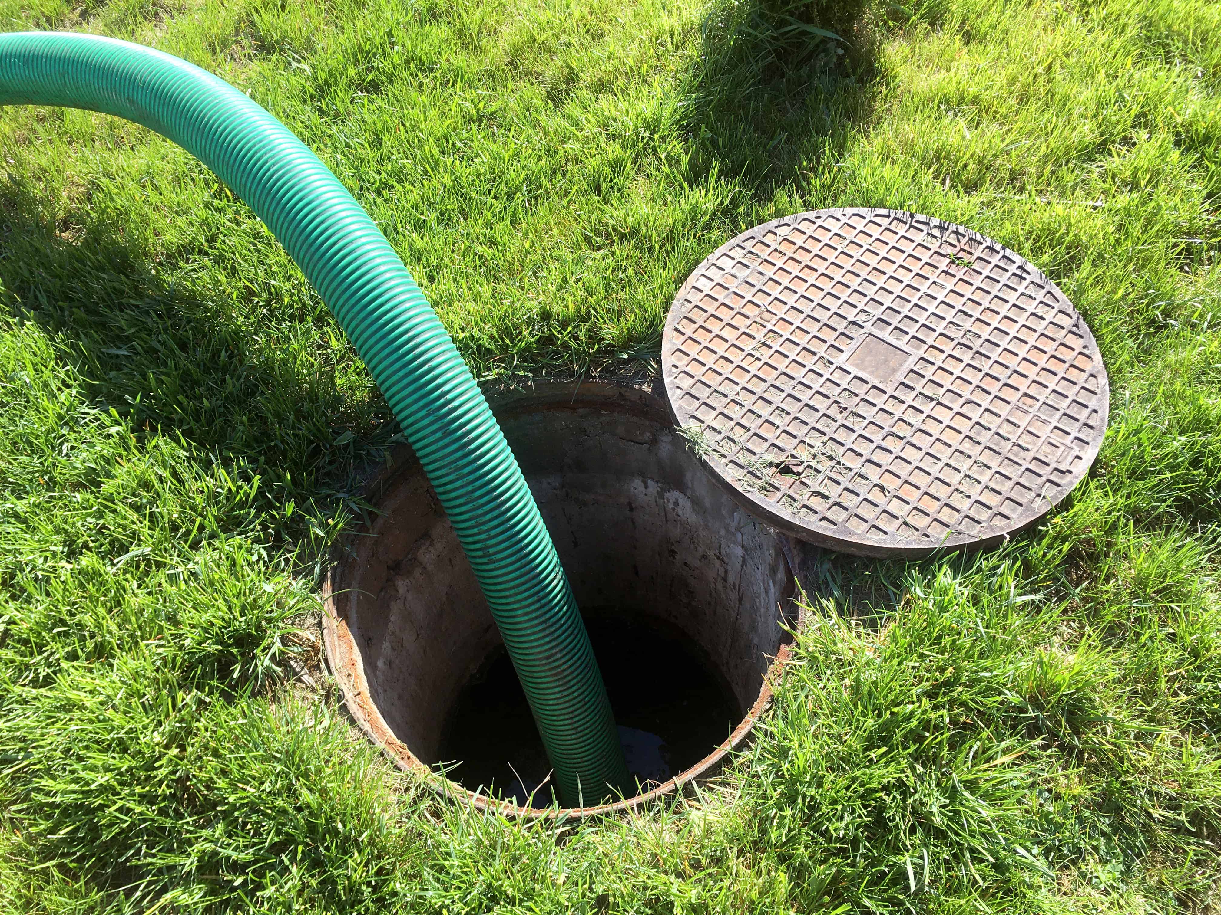 A green hose is inserted into an open manhole on a neatly trimmed lawn, with the circular cover resting to the side. The manhole appears to lead to a septic tank, suggesting it’s servicing for Septic Tank Pumping or Cesspool Cleaning.
