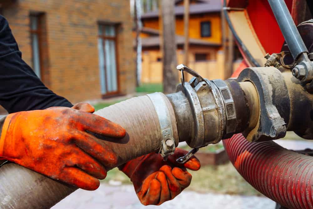 A person wearing bright orange gloves connects a large hose to a coupling on heavy machinery outdoors.