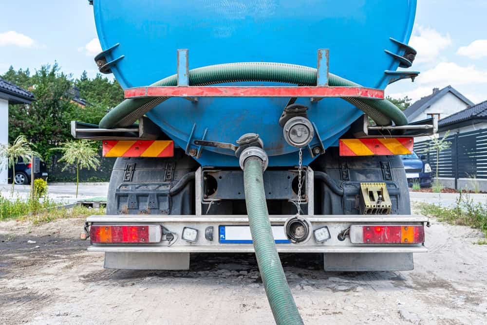A close-up rear view of a blue sewage or water tanker truck with hoses attached and hanging down, parked on a dirt surface in a residential area, providing septic tank services Suffolk County, NY.