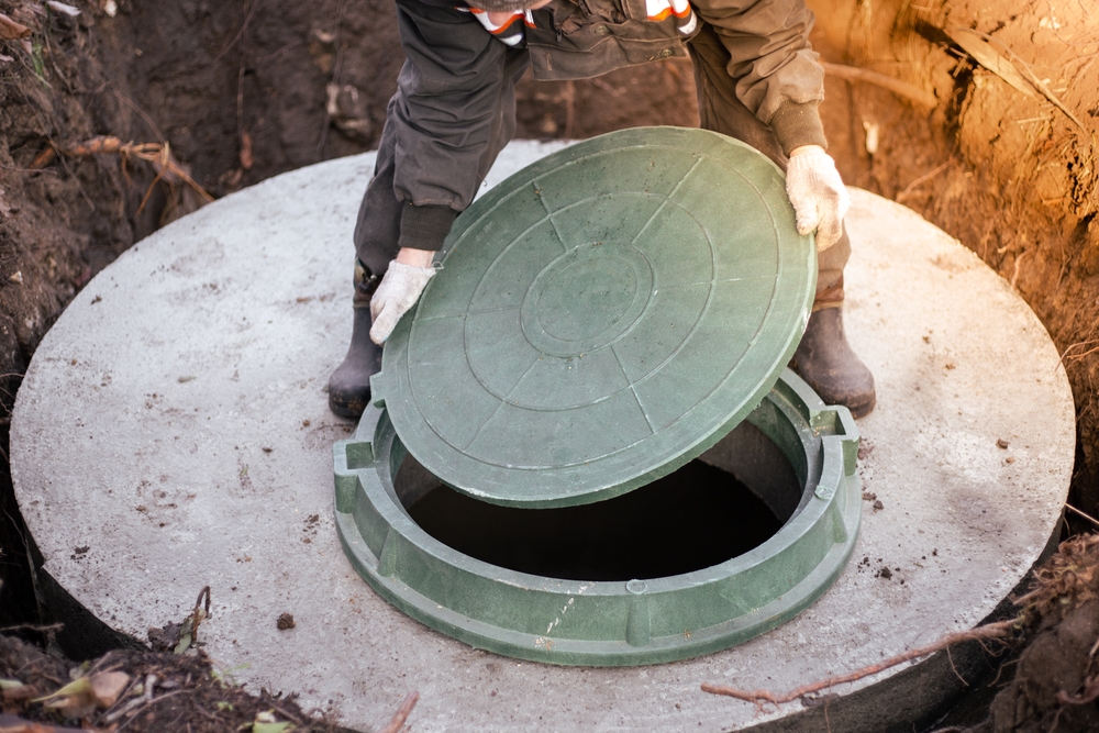 A person wearing gloves and work clothes lifts the green lid of a concrete outdoor septic tank or utility access hatch surrounded by soil, typical for cesspool services Suffolk County, NY.