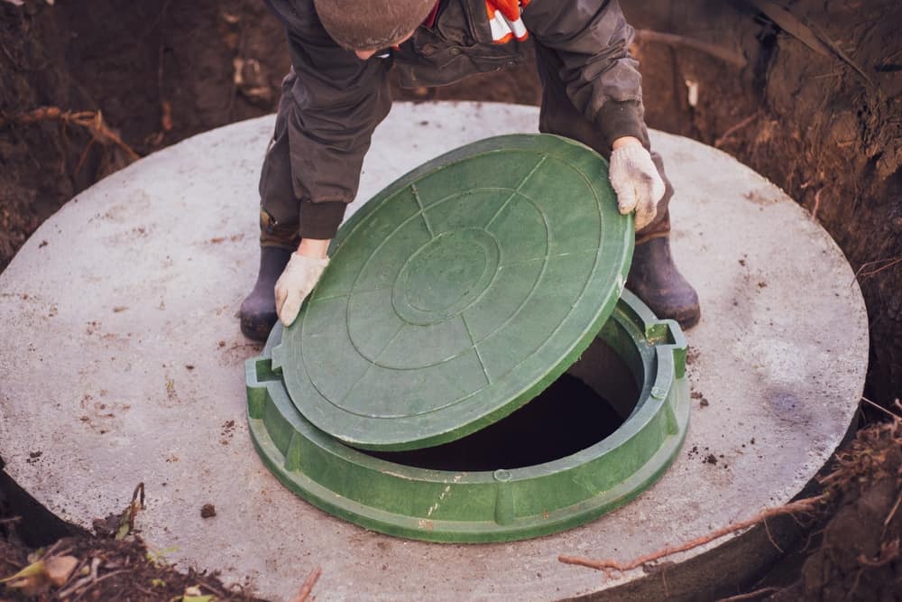 A person in work clothes and gloves opens a green circular manhole cover on a concrete surface, revealing an underground space.