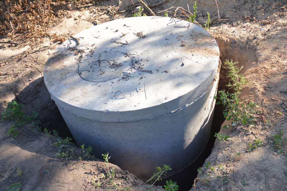 A large concrete septic tank is partially buried in the ground, surrounded by dirt and sparse green plants.