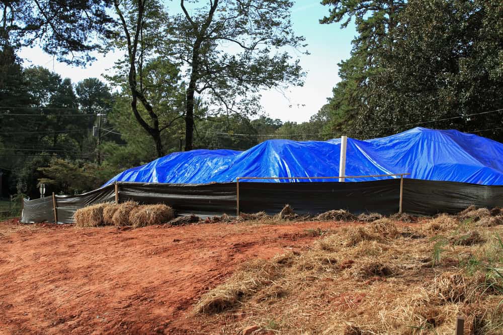 A construction site with a ground covered in red dirt and surrounding straw features a structure being built under large blue tarps, with a black barrier along the perimeter.