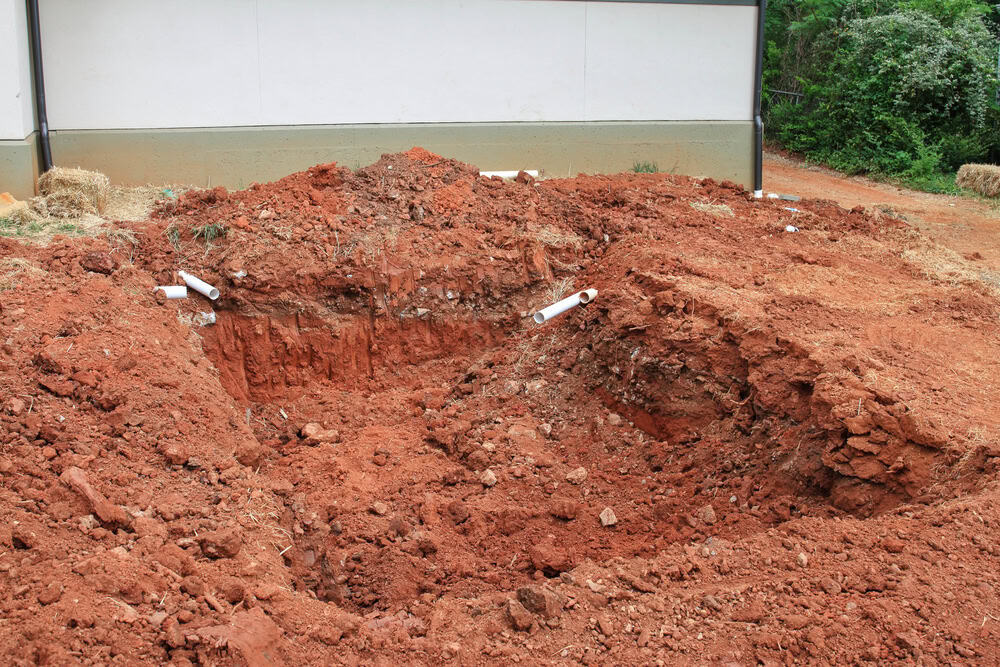 A large excavated hole with red soil in front of a house, featuring exposed white PVC pipes protruding from the ground.