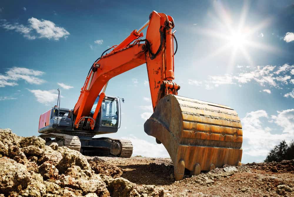 A large orange excavator with a metal bucket is digging into rocky soil under a bright blue sky with scattered clouds.