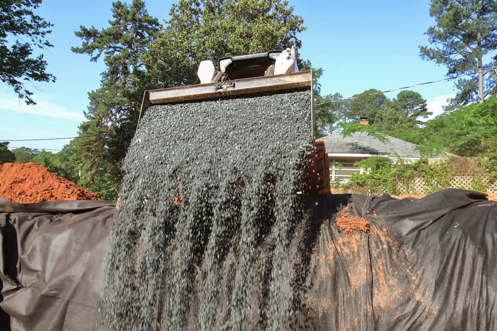 A construction scene where gravel is being poured from a large metal bucket onto a black tarp.