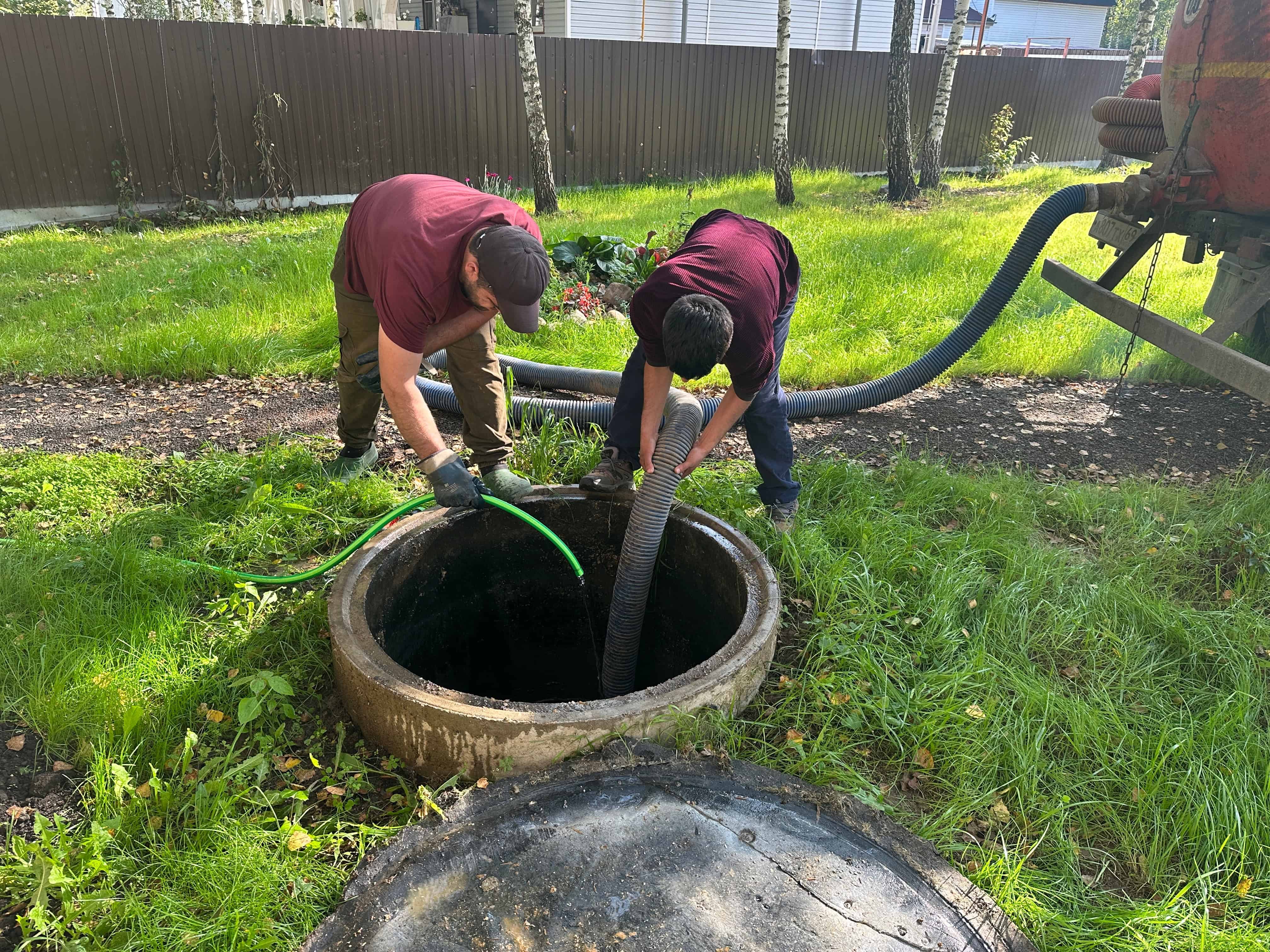 Two workers in maroon shirts and gloves are standing on grass, working on a sewer manhole. They are positioning hoses into the open manhole. The area is grassy with trees and a fence, indicating a typical scene for Septic Tank Services Suffolk County.