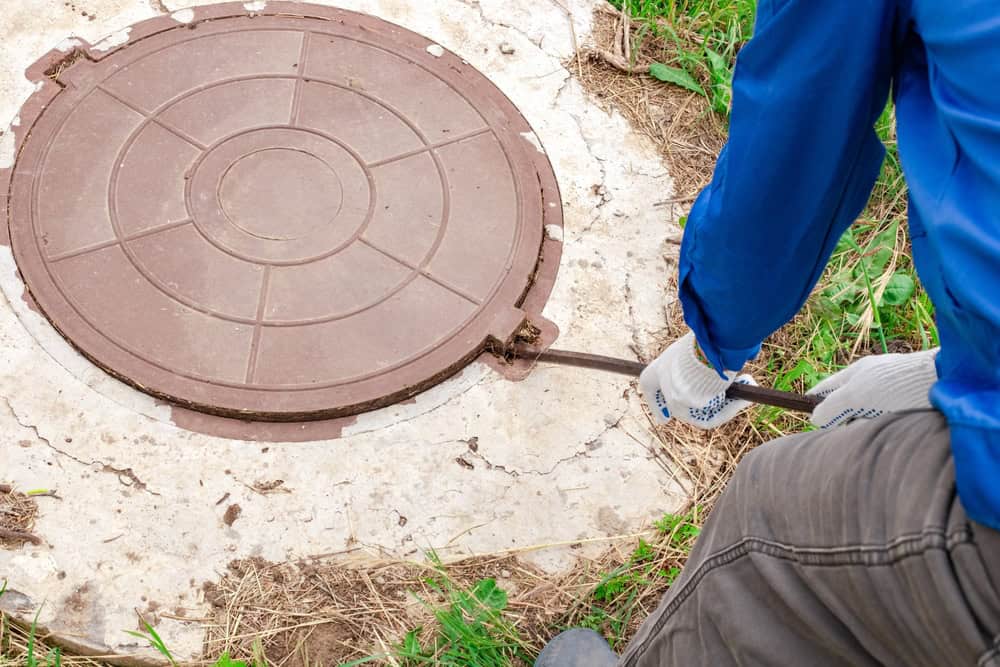 A person wearing blue clothing and white gloves is lifting a brown manhole cover with a metal tool. The manhole cover, set in a concrete surface surrounded by grass, appears to be part of routine cesspool services in Suffolk County.