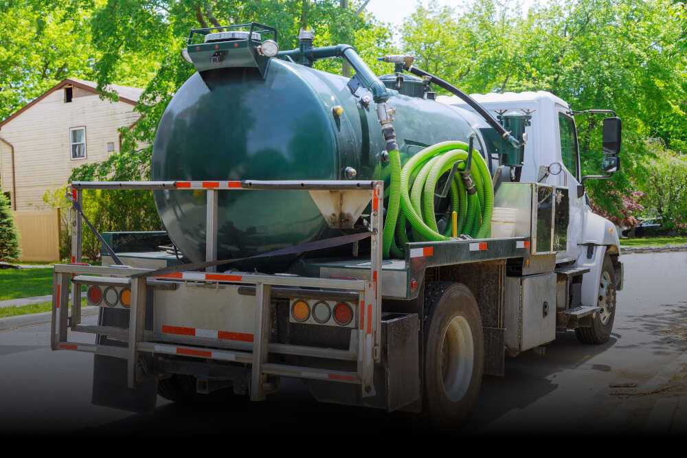 A large white vacuum truck with a green tank and green hoses, set for cesspool cleaning, is parked on a residential street.