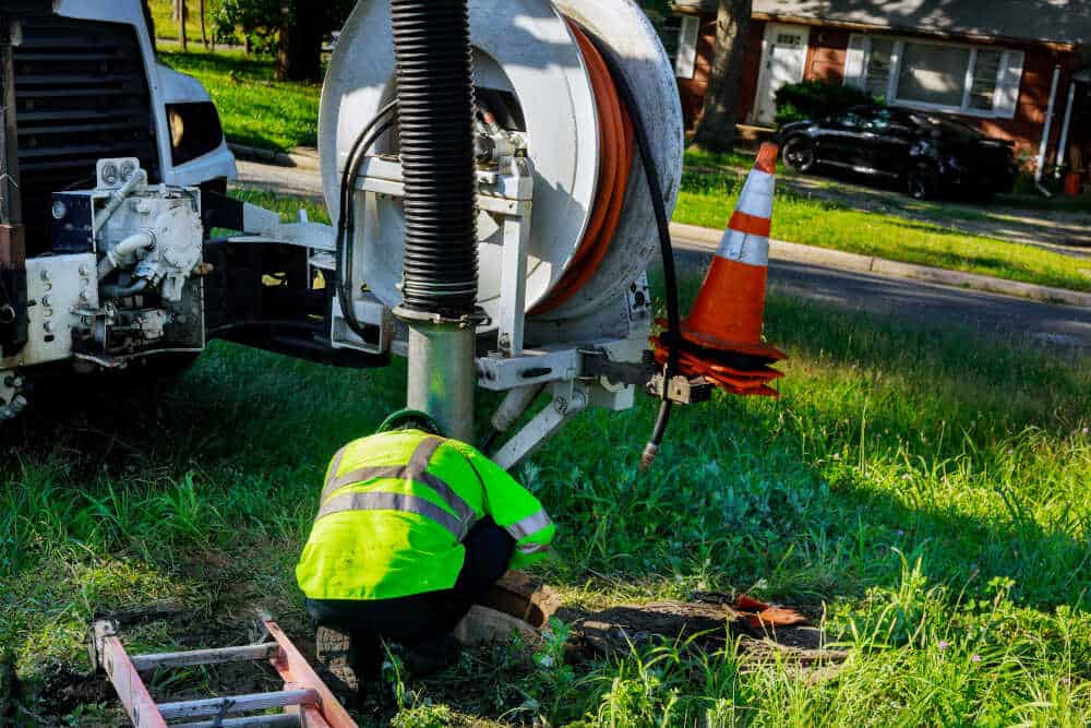 A worker in a bright green safety jacket bends over to inspect equipment attached to a large truck, possibly for septic tank pumping. Nearby, two orange safety cones are stacked, and a ladder lies on the grass. In the background, a residential street with a house and parked car is visible.