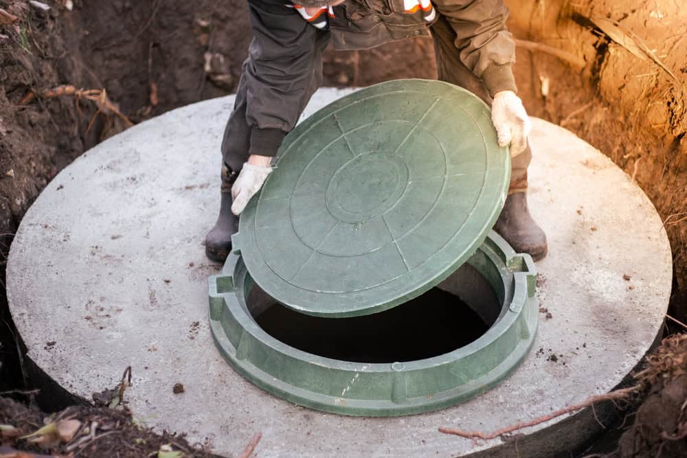 A person wearing gloves opens the green lid of a concrete septic tank outdoors in NY, revealing a dark opening—an essential step in professional septic tank services Suffolk County residents rely on.