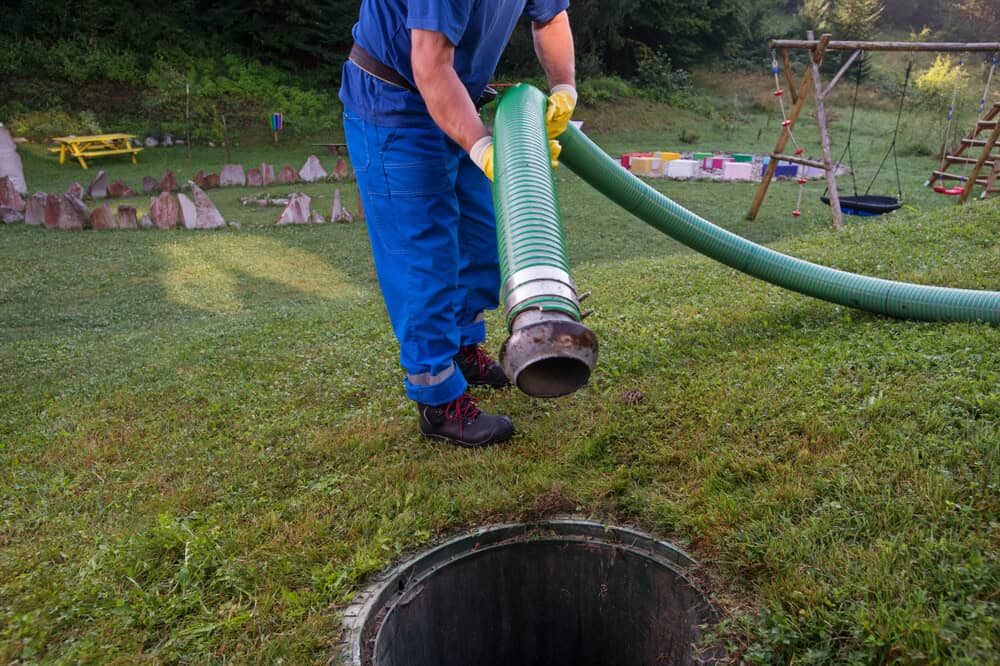 A worker in blue overalls holds a large green hose above an open septic tank in a grassy backyard, preparing for cleaning.