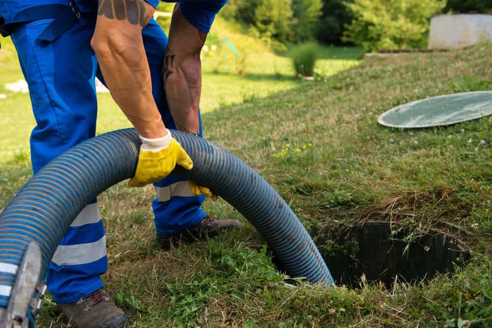 A worker in blue overalls and yellow gloves uses a large hose to clean or pump out a septic tank in a grassy NY yard; the septic tank lid is removed nearby, showcasing cesspool services Suffolk County, NY.