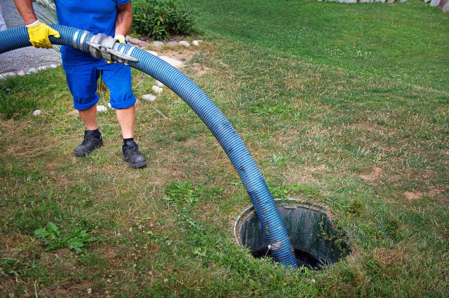 A person in blue work clothes and yellow gloves holds a large blue hose, placing it into an open outdoor septic tank on a grassy lawn, showcasing professional septic tank services Suffolk County, NY.
