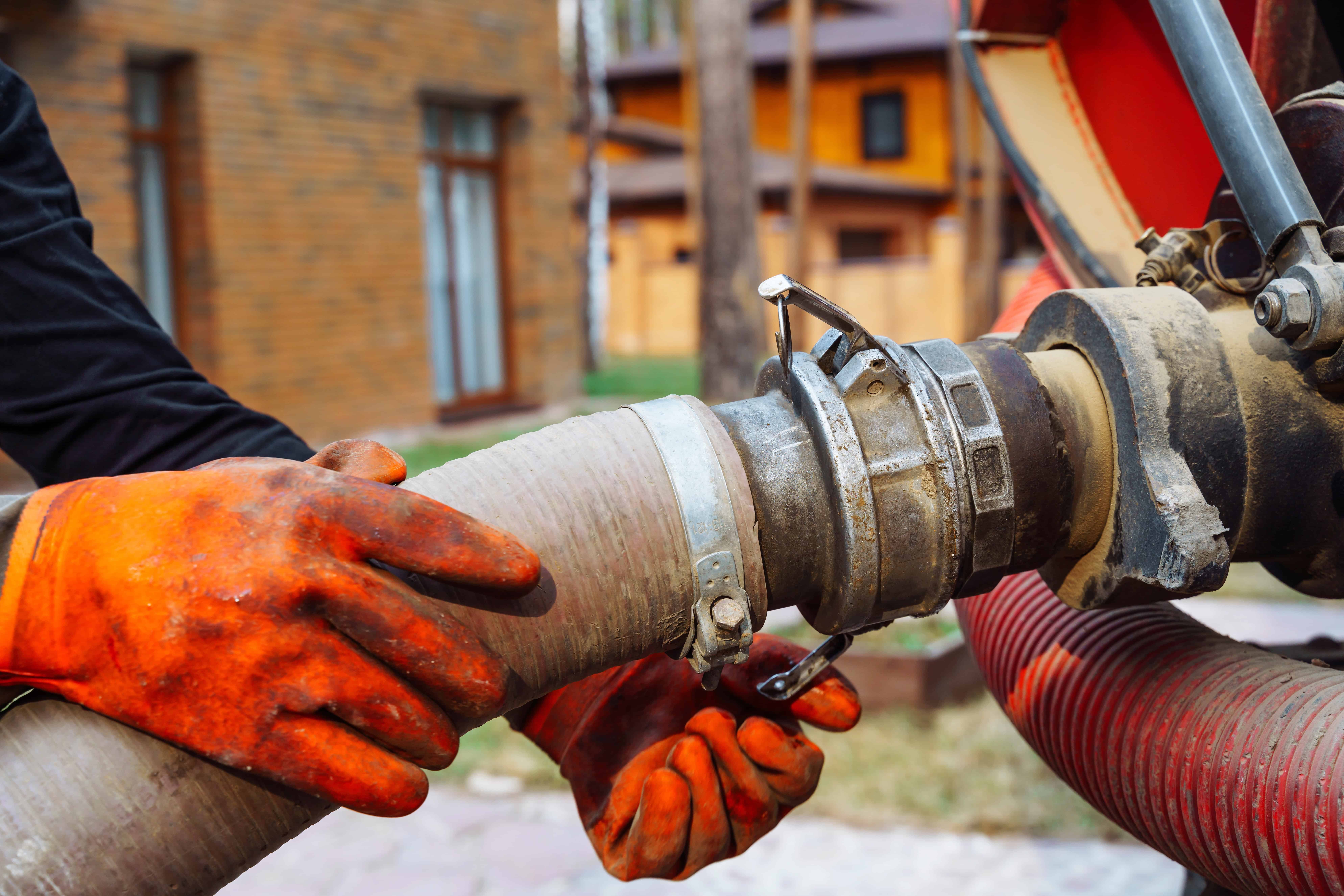 A person in orange gloves is connecting a large hose to industrial equipment for septic tank pumping outdoors. In the background, there are two wooden houses, trees, and other residential surroundings.