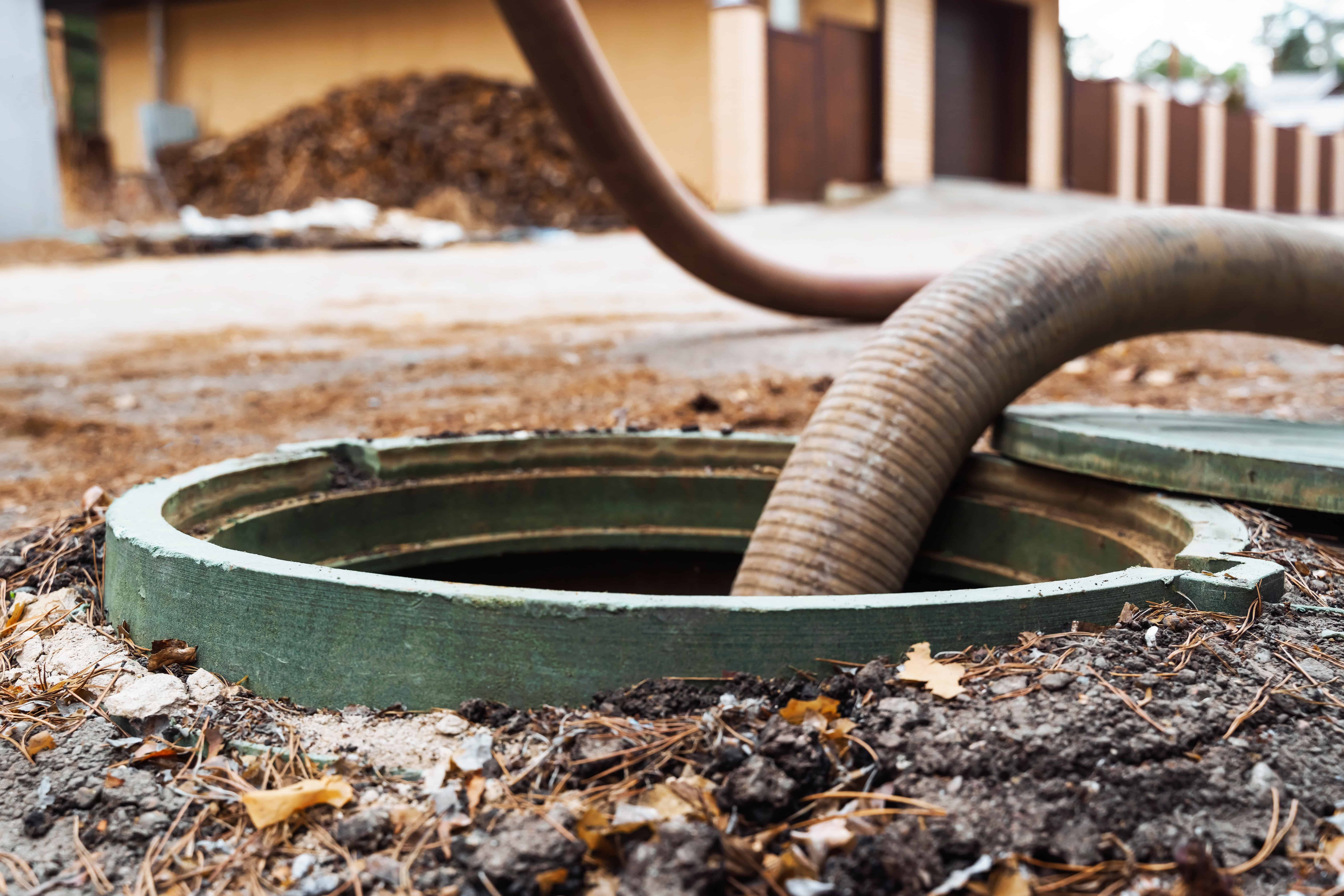 A large hose is inserted into an open green manhole on a dirt ground outside a building, suggesting septic tank pumping is in progress. The scene appears to be involved in maintenance or cleaning work, with the manhole cover partially visible and some debris scattered around.