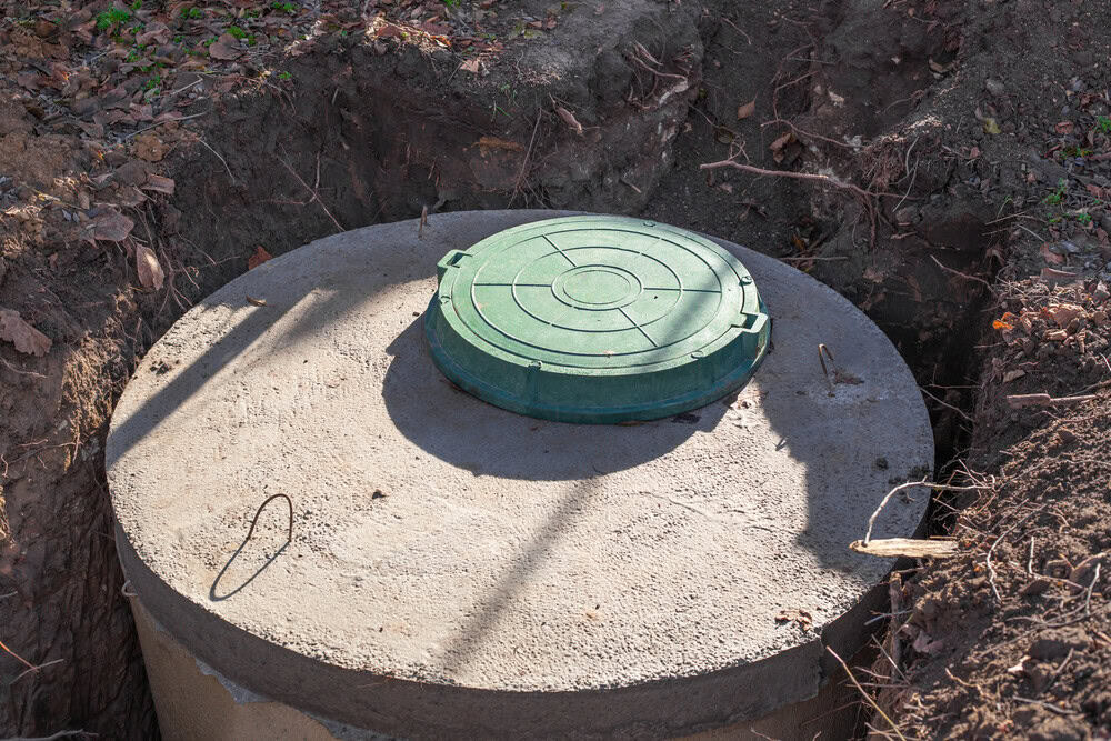 A concrete septic tank, partially buried in the ground with a green plastic access lid, is surrounded by exposed soil and fallen leaves.