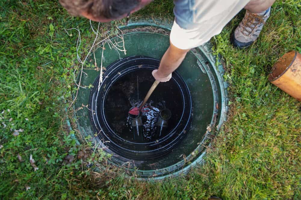 A person stands next to an open septic tank, using a tool to inspect or clean inside. The grassy area hints at residential NY surroundings—ideal for those seeking professional septic tank services Suffolk County. Only part of the person's shoes and arm are visible.