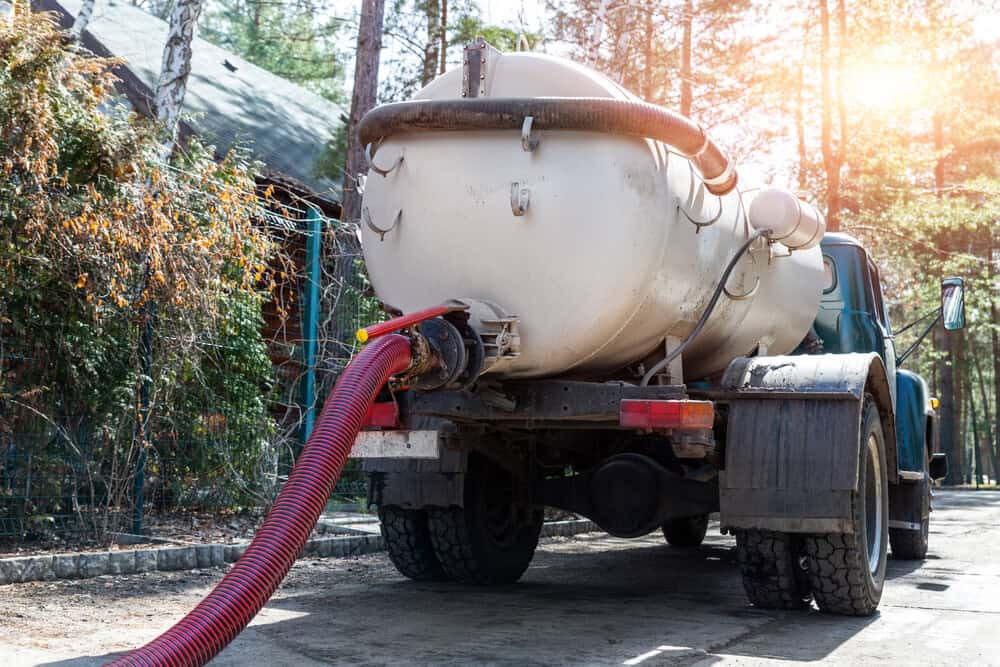 A vacuum truck with a large tank and a red hose is parked on a driveway in a wooded area, likely providing cesspool services Suffolk County, NY, near a house. Sunlight filters through the surrounding trees.