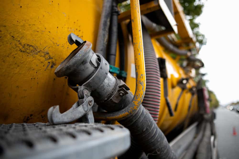 Close-up of industrial equipment featuring a yellow, tank-like structure used for cesspool cleaning, with various attached hoses and pipes. A metallic component with a handle is prominently visible in the foreground. The background is slightly blurred, showing trees and an overcast sky.