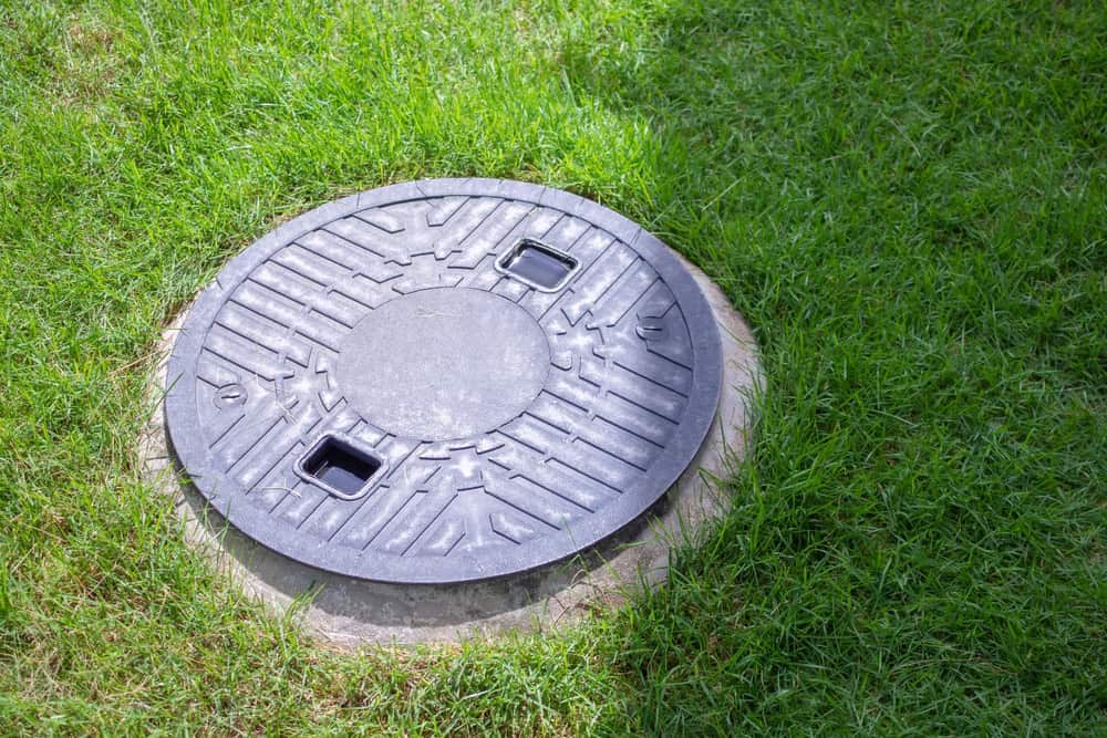 A round, black manhole cover with two small rectangular slots is situated on a grassy lawn, likely indicating a point for septic tank pumping. The cover has a textured, geometric pattern and is slightly raised from the surrounding grass.