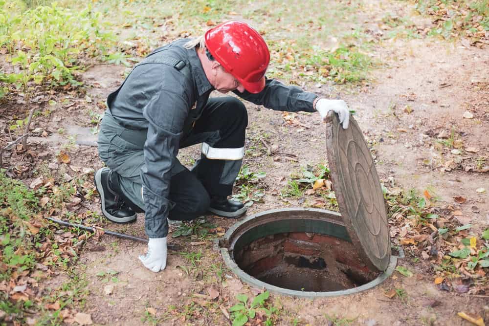 A worker wearing a red hard hat, gray overalls, and white gloves is kneeling next to an open manhole cover in an outdoor setting. He appears to be inspecting or accessing the cesspool. Fallen leaves and greenery are visible on the ground around him.