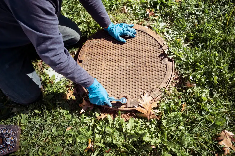 A person wearing blue gloves kneels on grass and uses a wrench to work on a round manhole cover surrounded by fallen leaves.