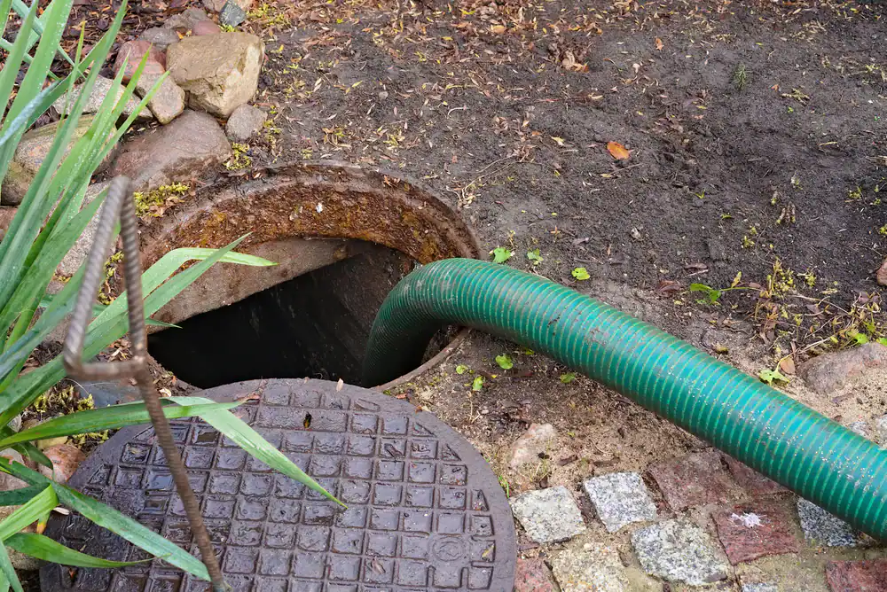 A green hose is inserted into an open, rusty manhole surrounded by dirt, grass, and stone pavers, with the metal cover lying nearby on the ground.