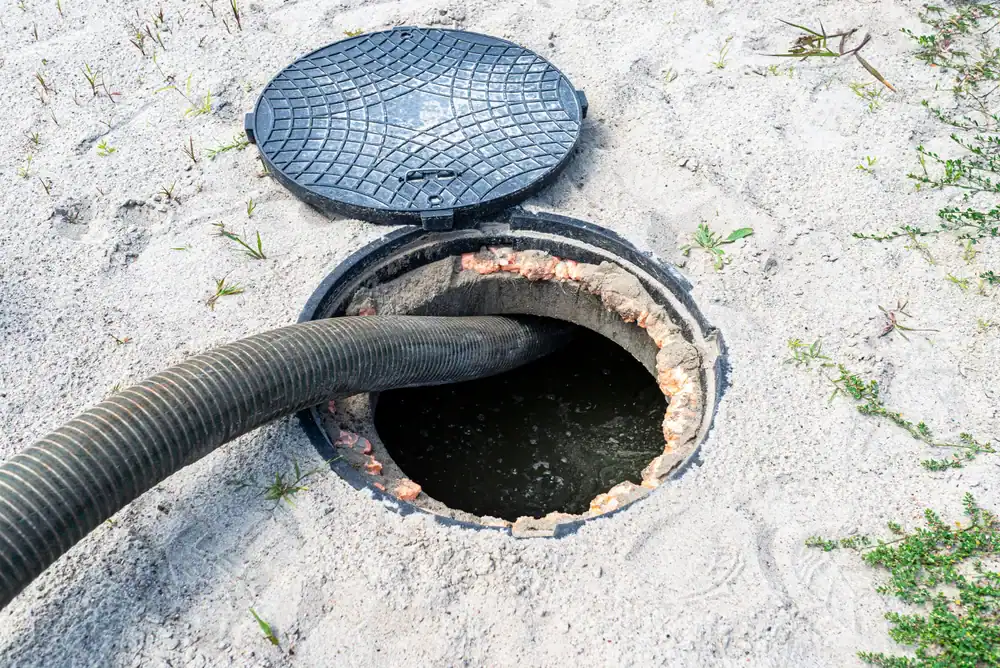 A large black hose inserted into an open septic tank in sandy soil, with the round manhole cover resting to the side and some grass growing nearby.