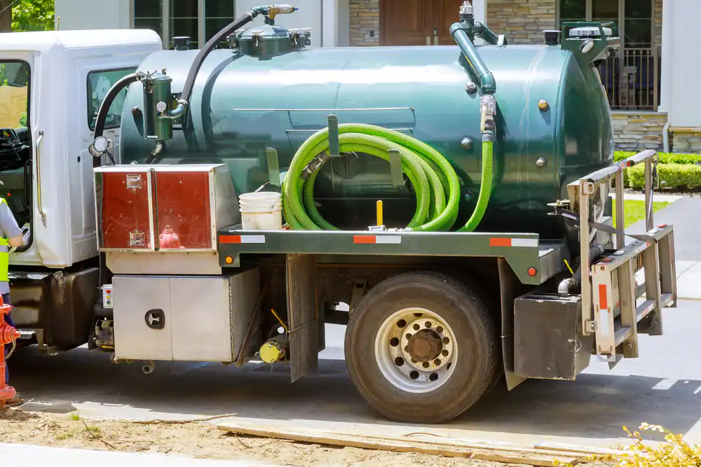 A green and silver vacuum truck with hoses and tanks is parked on a residential street, next to a sidewalk and some grass.