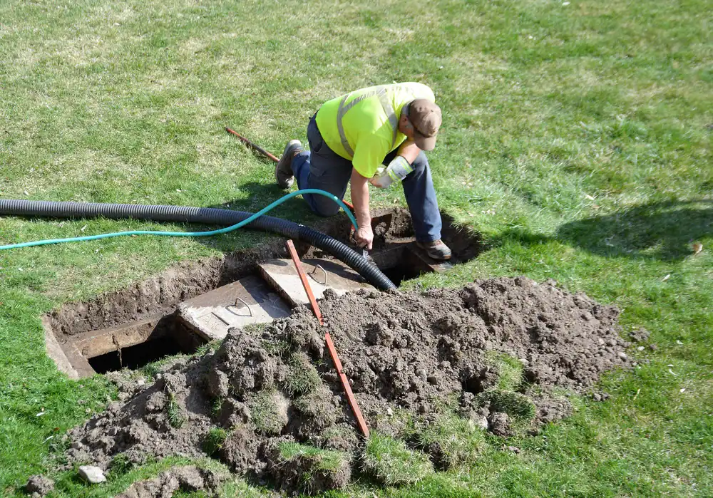 A worker in a yellow safety vest kneels on grass while installing or inspecting black tubing in a dug trench with exposed soil and tools beside an open rectangular ground cover.