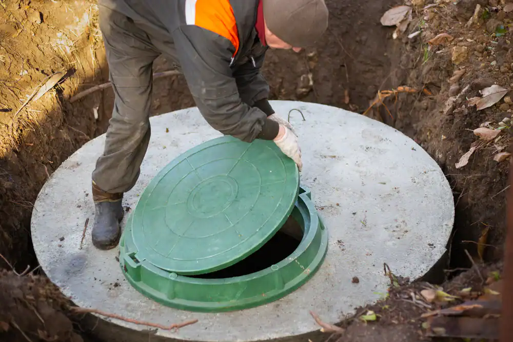 A person in work clothes and gloves is lifting the green lid of a concrete septic tank or underground access point. The person is standing in a pit surrounded by dirt and fallen leaves.