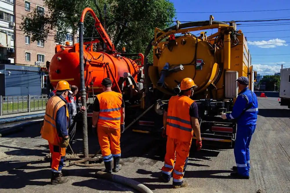 Five construction workers in safety gear operate equipment between two large industrial vehicles on a sunny street. Some workers handle hoses, while one in blue takes notes. Buildings and trees are visible in the background.