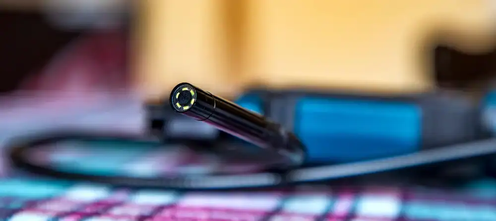 A close-up of an endoscope camera with a flexible black tube, lying on a checkered surface, with a blurred blue object and yellow background.