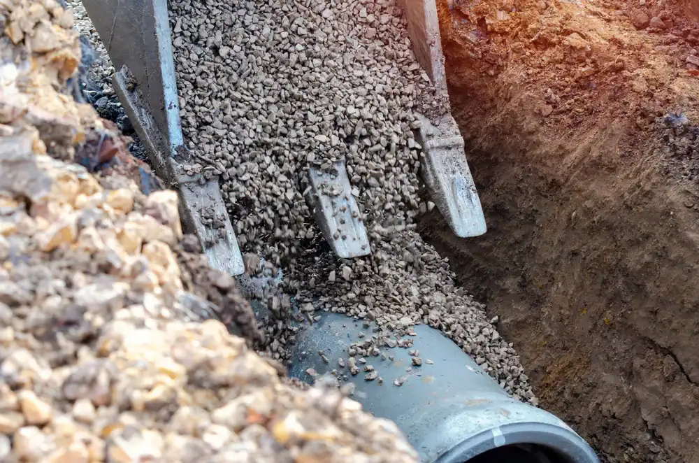 Excavator bucket pouring gravel over a large gray drainage pipe in a trench at a construction site, preparing for pipe installation and ground covering.