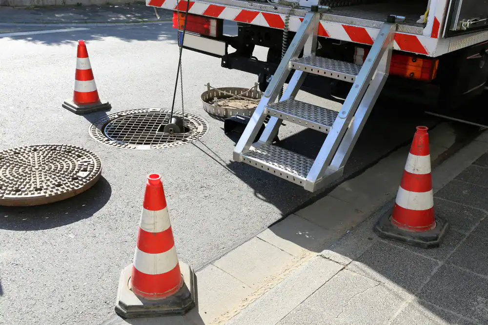 A utility truck is parked by an open manhole on a street. Safety cones surround the area, and a metal ladder extends from the truck toward the sidewalk. The manhole cover is removed and lies on the ground nearby.